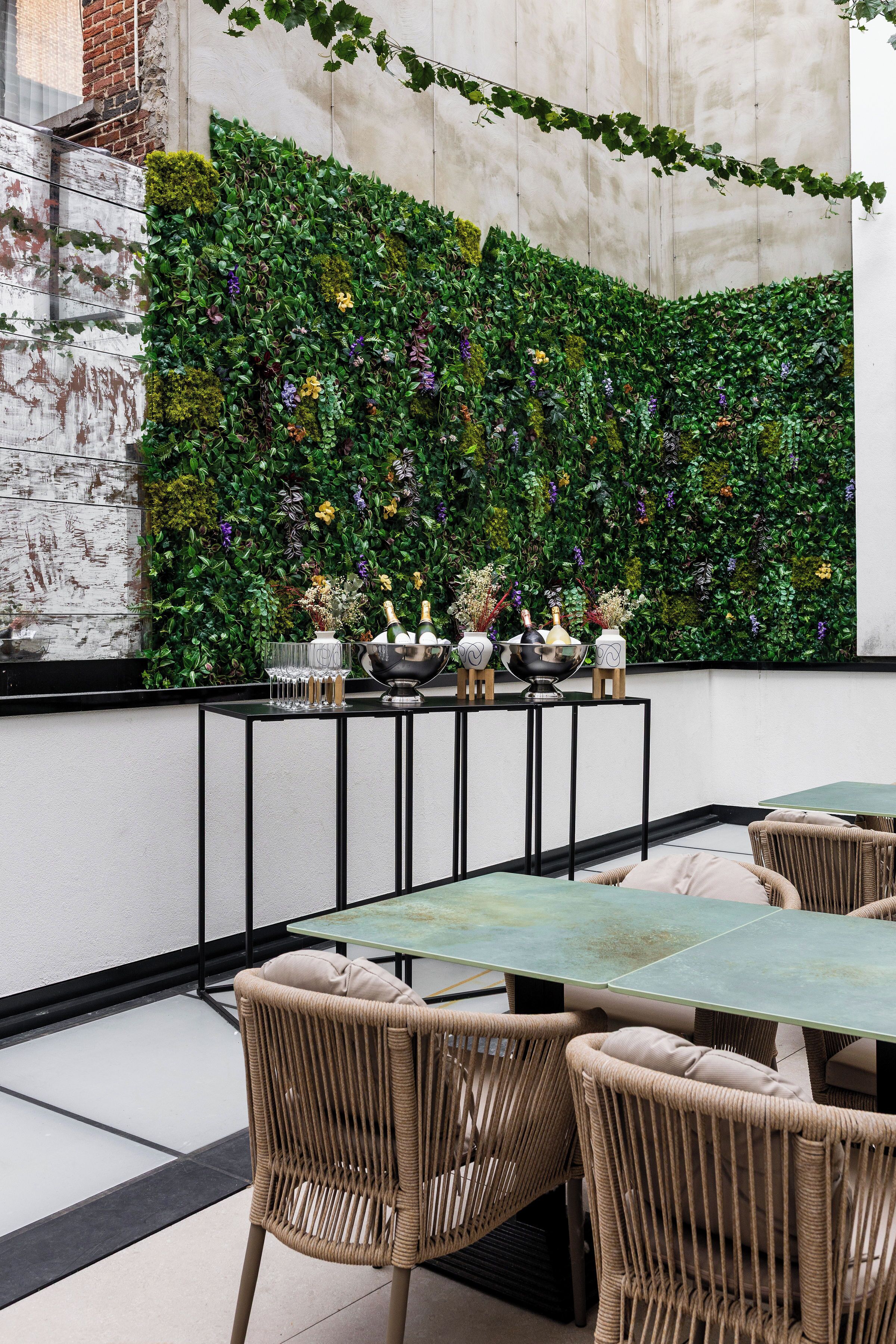 Un espacio de esparcimiento al aire libre con sillas de mimbre y una mesa, una pared verde con plantas y una zona de bar con vasos y botellas.