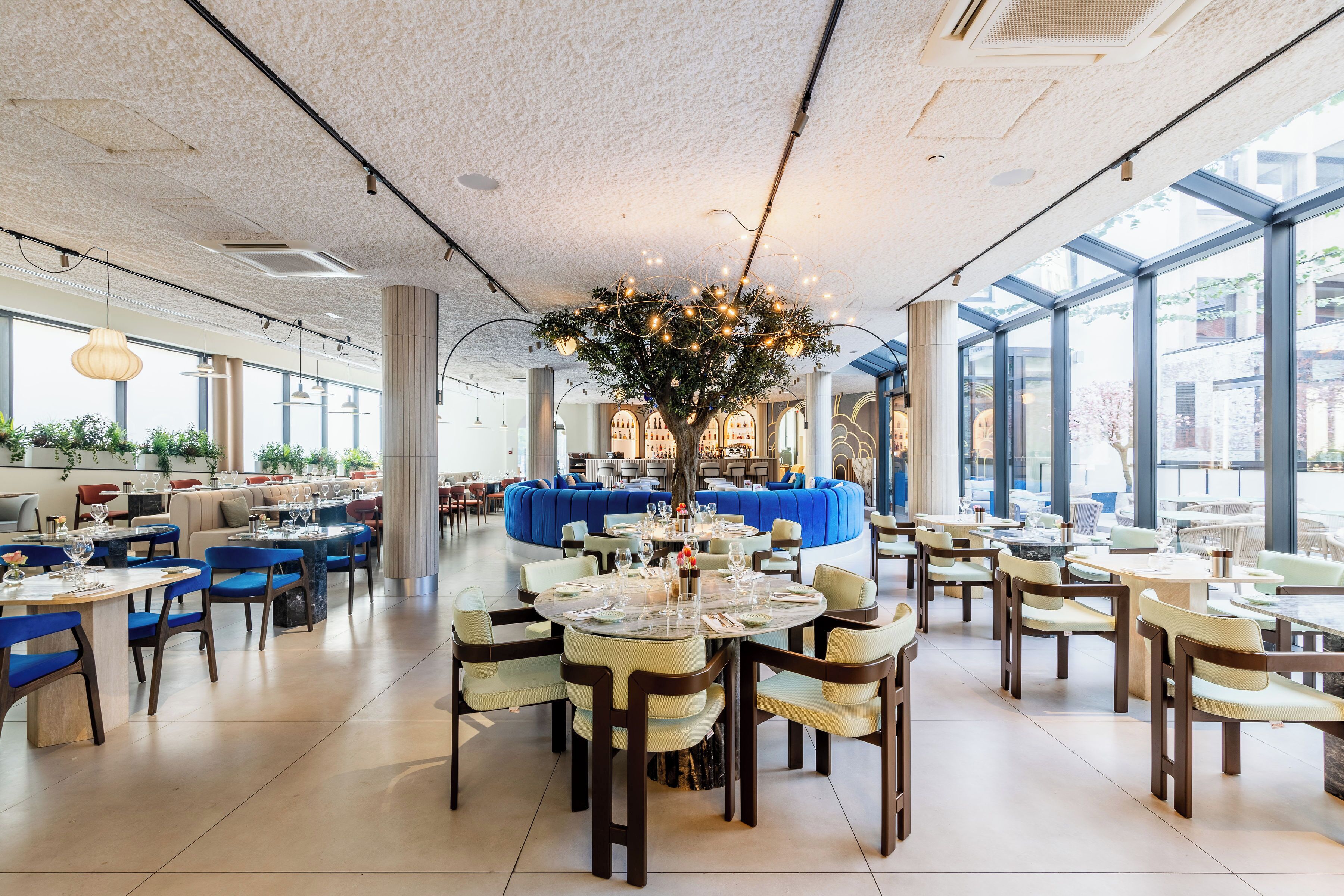 A spacious restaurant with round tables, blue seating, and a central decorative tree.