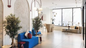 A modern interior with a blue sofa, two potted plants, a round coffee table, and a large window with natural light. - The Liman Hotel (Brussels)