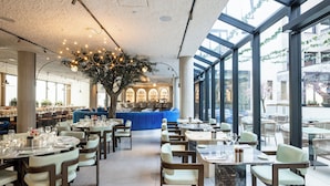 A restaurant with tables, chairs, a large tree, and a view through large windows. - The Liman Hotel (Brussels)