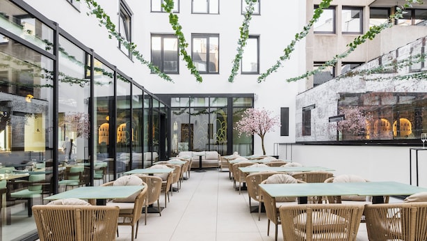 A modern indoor dining area with glass walls, wicker furniture, and a central seating arrangement. - The Liman Hotel (Brussels)