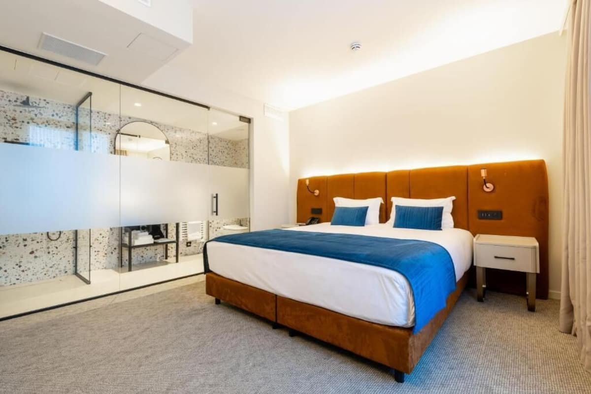 A modern hotel room with a large bed, a glass-enclosed bathroom, and a wooden headboard.