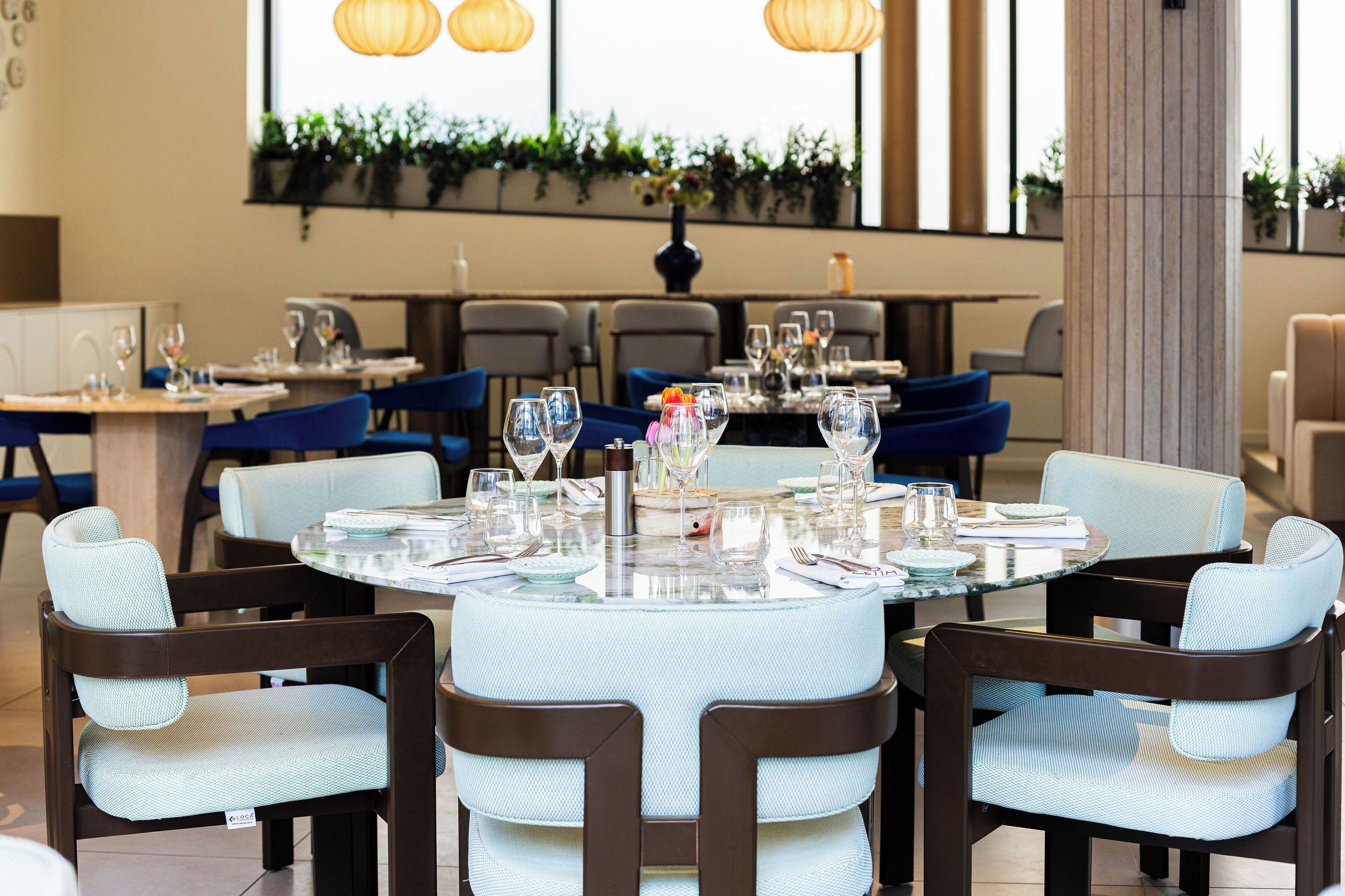 A restaurant with round tables, chairs, and a large window with plants.