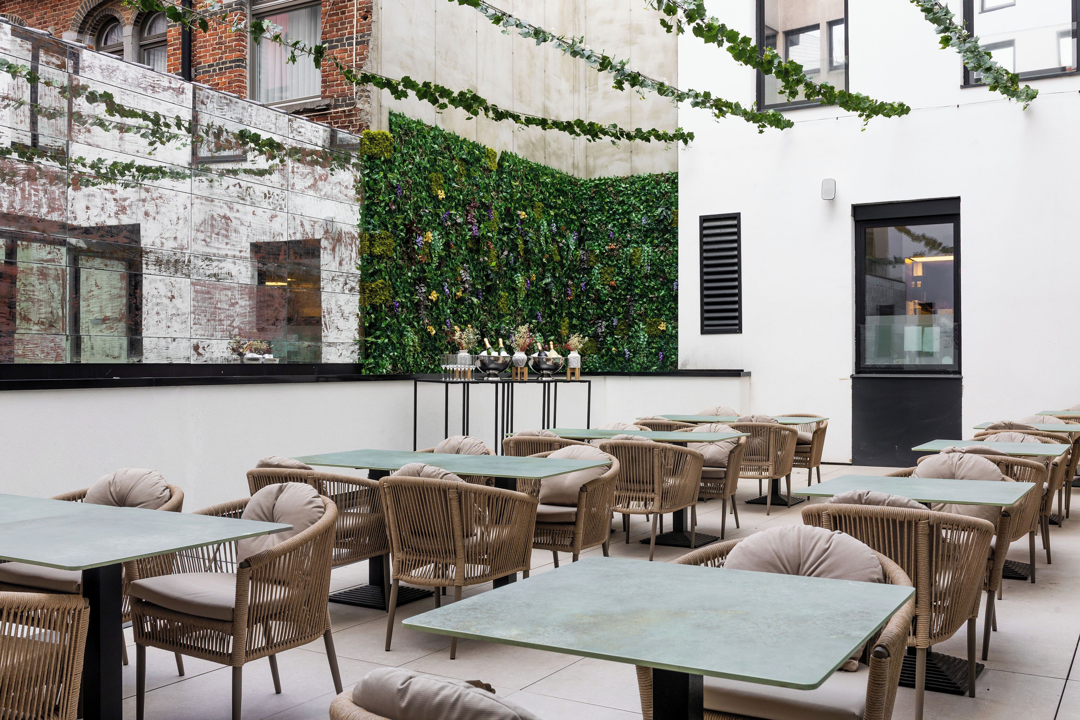 An outdoor dining area with wicker chairs and tables, a brick wall with greenery, and a modern building in the background.