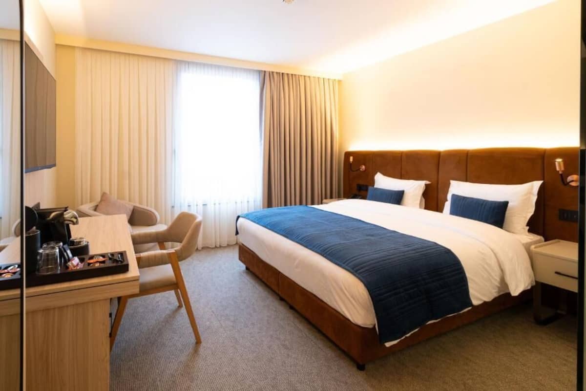 A hotel room with a large bed, a desk with a chair, a dresser, and a window with curtains.