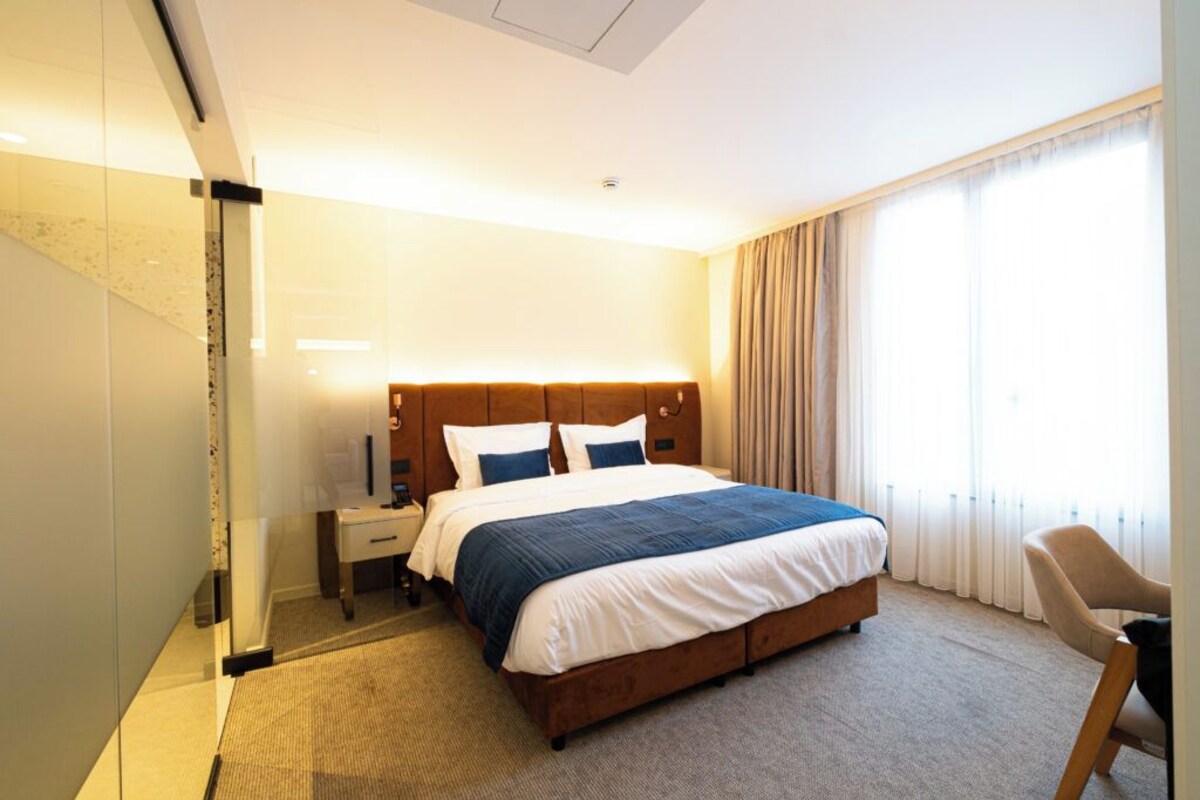 A hotel room with a bed, bedside table, chair, and a large window with curtains.