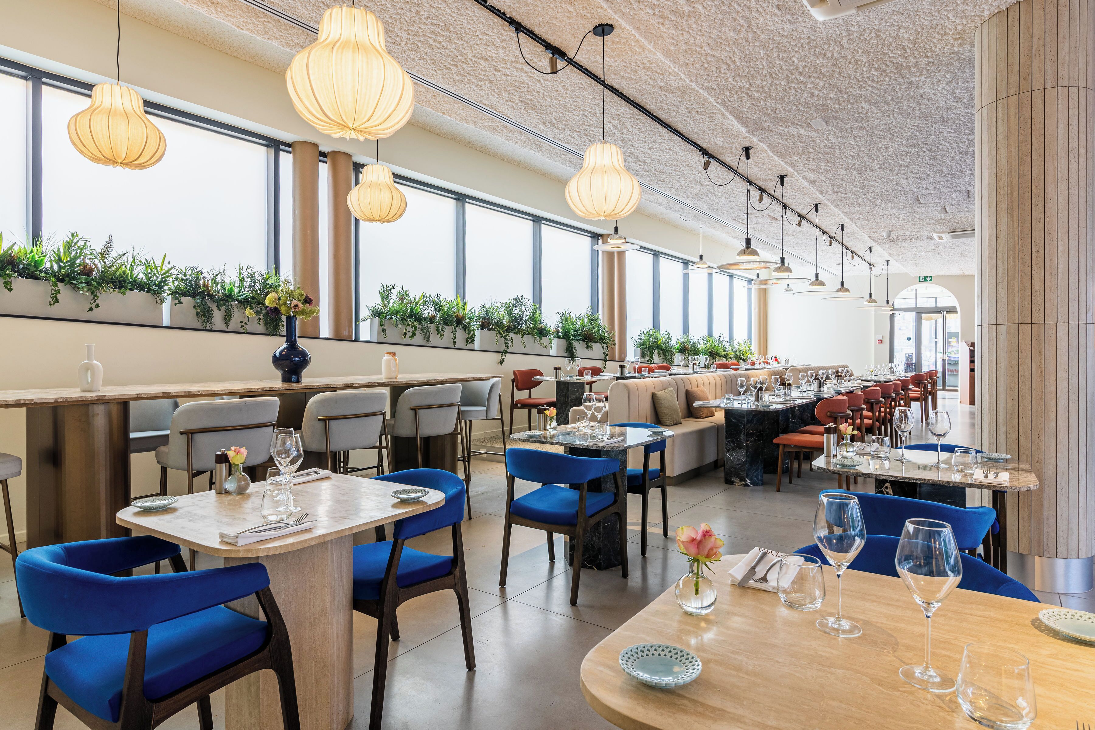 Een restaurant met meerdere eettafels en stoelen, grote ramen, hangende吊灯 en een wand vol planten.
