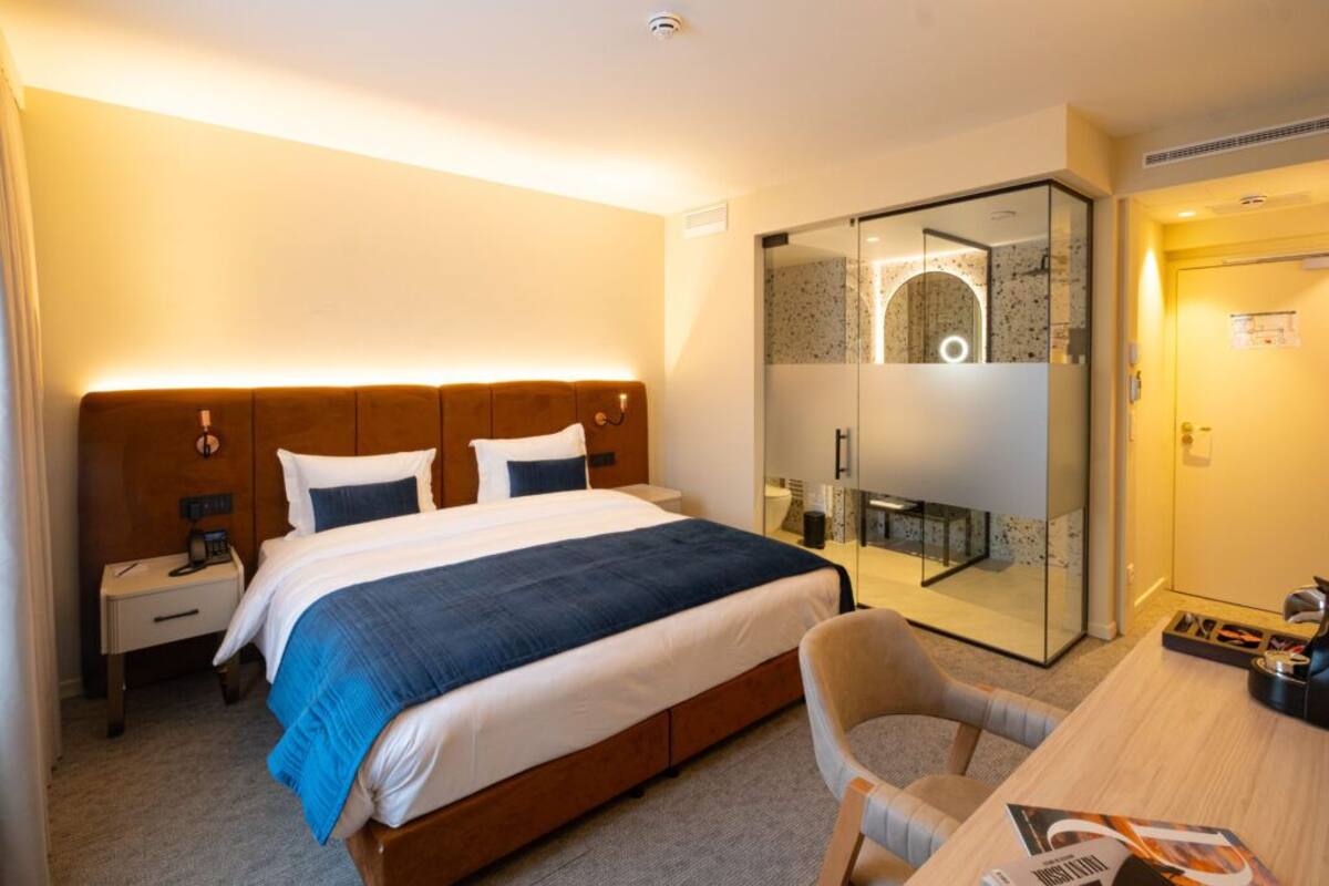 A hotel room with a bed, bedside table, chair, desk, and a glass-enclosed shower area.