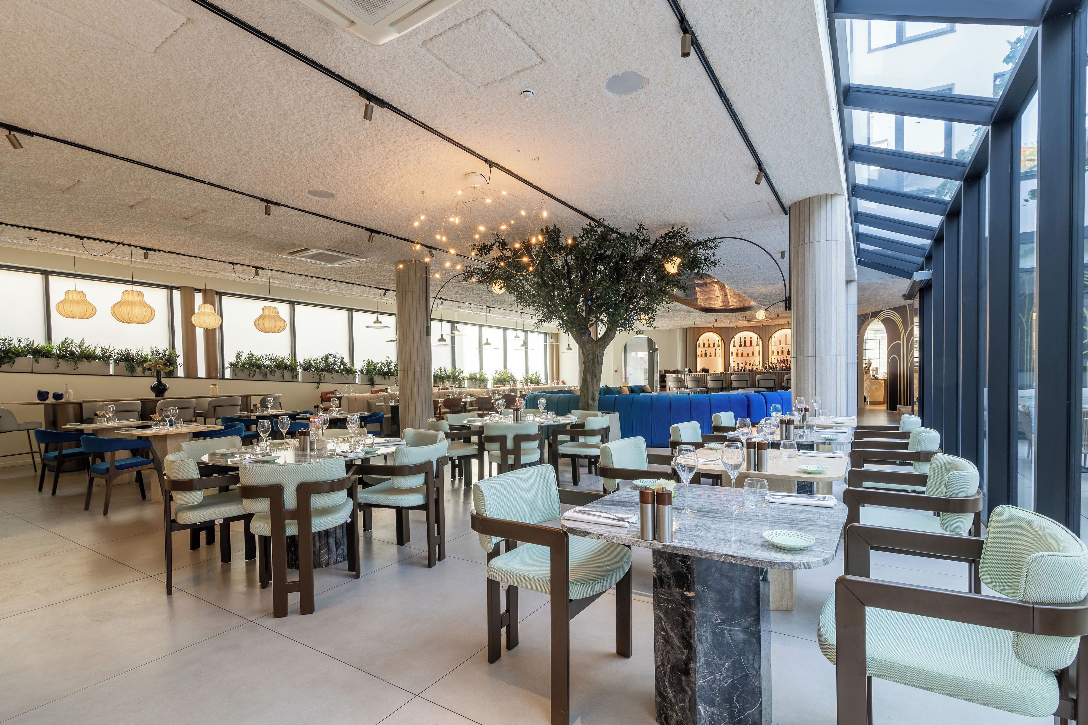 A spacious restaurant with high ceilings, large windows, and a central tree. Tables are arranged in a casual layout, with chairs and dining tables.