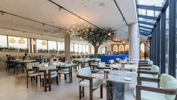 A spacious restaurant with high ceilings, large windows, and a central tree. Tables are arranged in a casual layout, with chairs and dining tables.