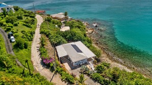 Ett flygbildsfoto av en fastighet vid kusten med ett hus, en väg och fri utsikt över havet.