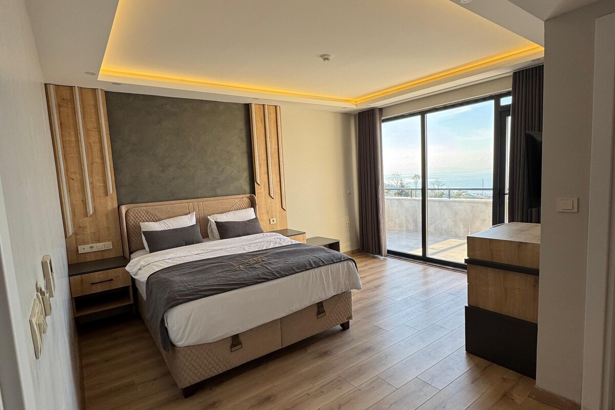 A modern bedroom with a large bed, wooden flooring, a view of the city, and a balcony.