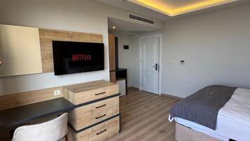 Moderni makuuhuone, jossa on puinen työpöytä, harmaalla peitolla varustettu sänky ja televisio, jonka näytöllä näkyy teksti ”NETFLIX”.
