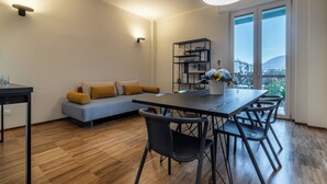 Una sala de estar moderna con un sofá, una mesa de comedor con sillas, una estantería y vistas de la ciudad a través de amplios ventanales.