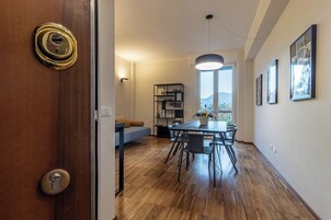 Un interior moderno con piso de madera, una zona de comedor con mesa y sillas, un sofá, una estantería y un ventanal con vistas al exterior.