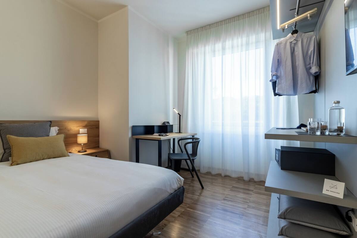 A hotel room with a bed, desk, chair, and a wardrobe with a 'No Smoking' sign.