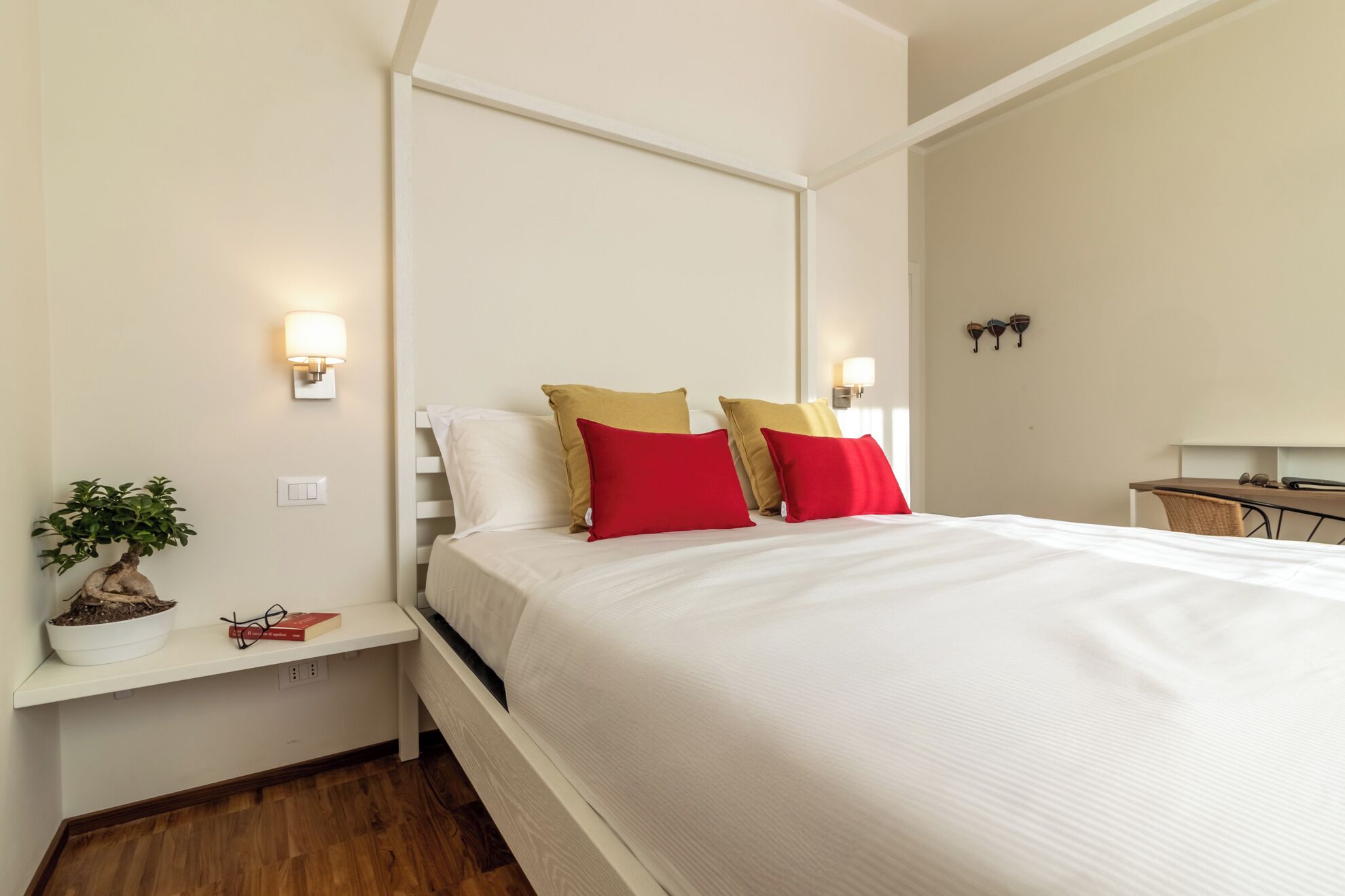 A neatly made bed with white and red pillows, a bedside table with a plant and book, and a wall-mounted lamp.