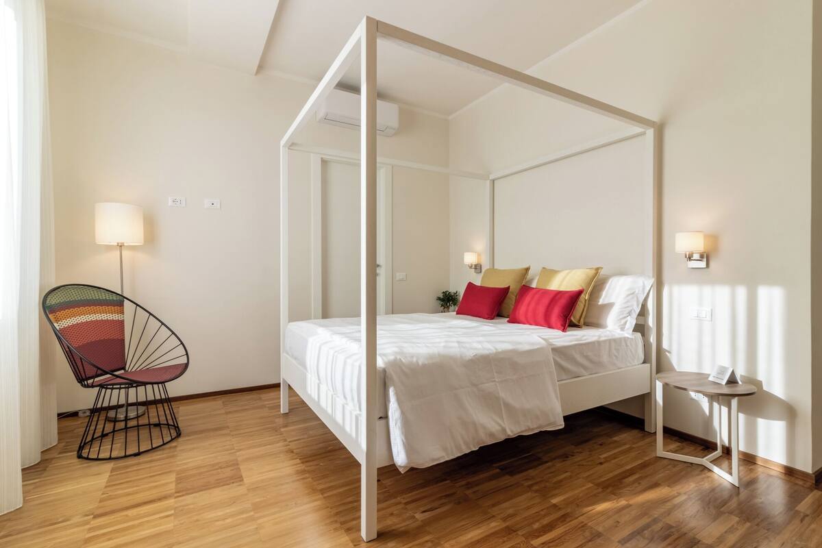 A four-poster bed with white bedding, a colorful chair, and a small bedside table with a lamp.