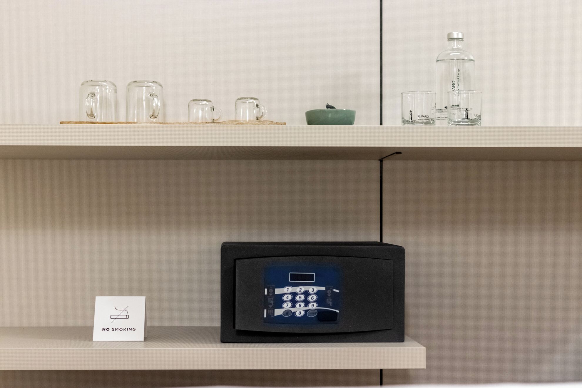 Una habitación con cartel de no fumar, una caja de seguridad digital y una estantería con cristalería y una botella.