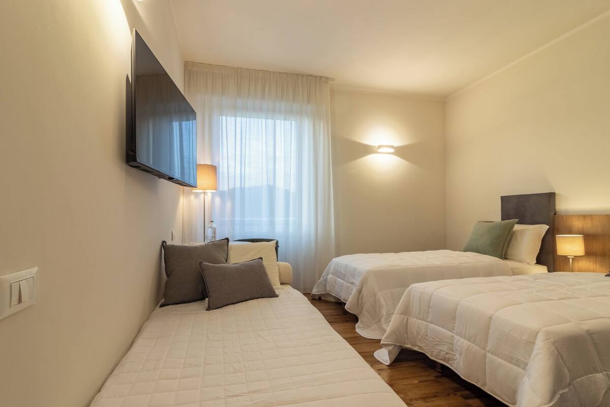 A hotel room with two beds, a TV mounted on the wall, a bedside table with a lamp, and a window with curtains.