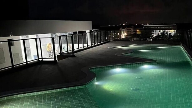 A nighttime swimming pool with a curved design, surrounded by a low wall and a building with glass doors. - Amazing apartment in Porto de Galinhas with stunning views close to the center (Porto de Galinhas Ipojuca)