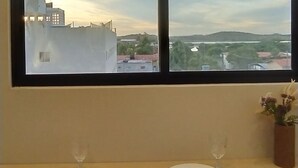 A hotel room with a large window offering a view of the cityscape and a table set with two wine glasses and plates. - Amazing apartment in Porto de Galinhas with stunning views close to the center (Porto de Galinhas Ipojuca)