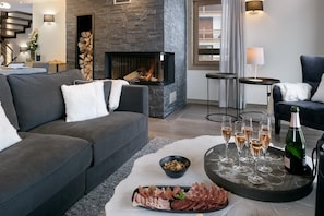 A modern living room with a fireplace, grey sofas, a tray with glasses and a bottle, and a small table with a lamp.