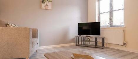 A living room with a sofa, a coffee table, a flat-screen TV on a stand, and a window with blinds.