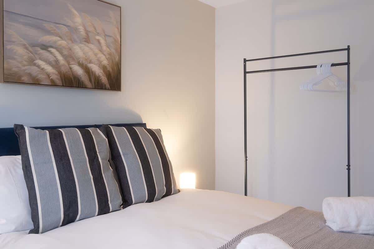Une chambre d’hôtel avec un lit, des coussins à rayures, un porte-vêtements et un tableau au mur.