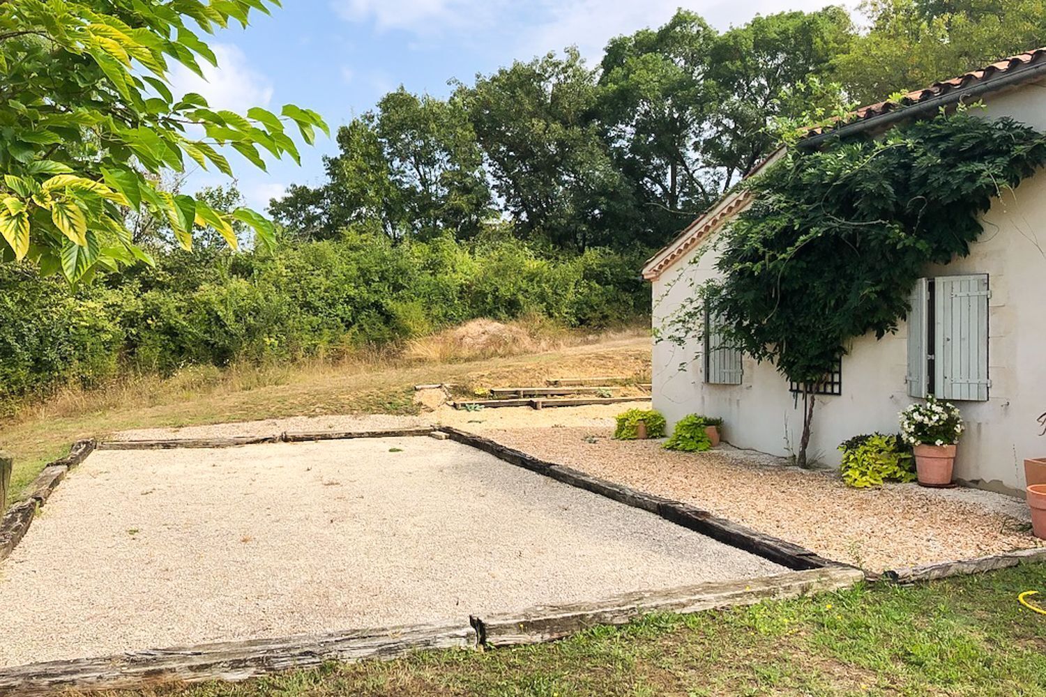 Un patio de grava con un cerramiento de madera, un edificio blanco con contraventanas y abundante vegetación circundante.