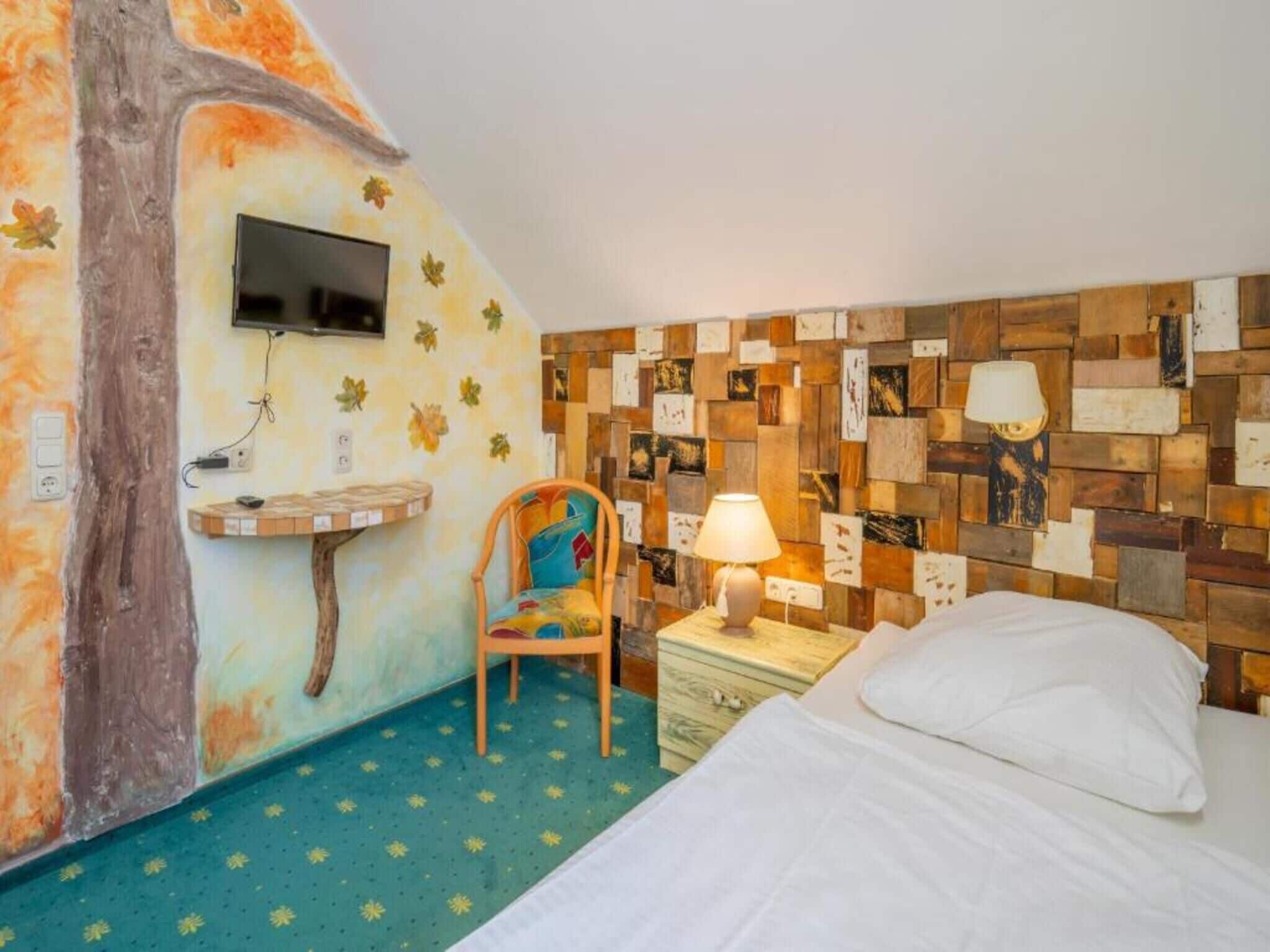 A room with a bed, a chair, a TV mounted on the wall, and a wall with a mosaic of wooden pieces.