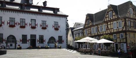 Una plaza adoquinada con edificios históricos, incluyendo un edificio blanco con adornos rojos y una estructura de entramado de madera.