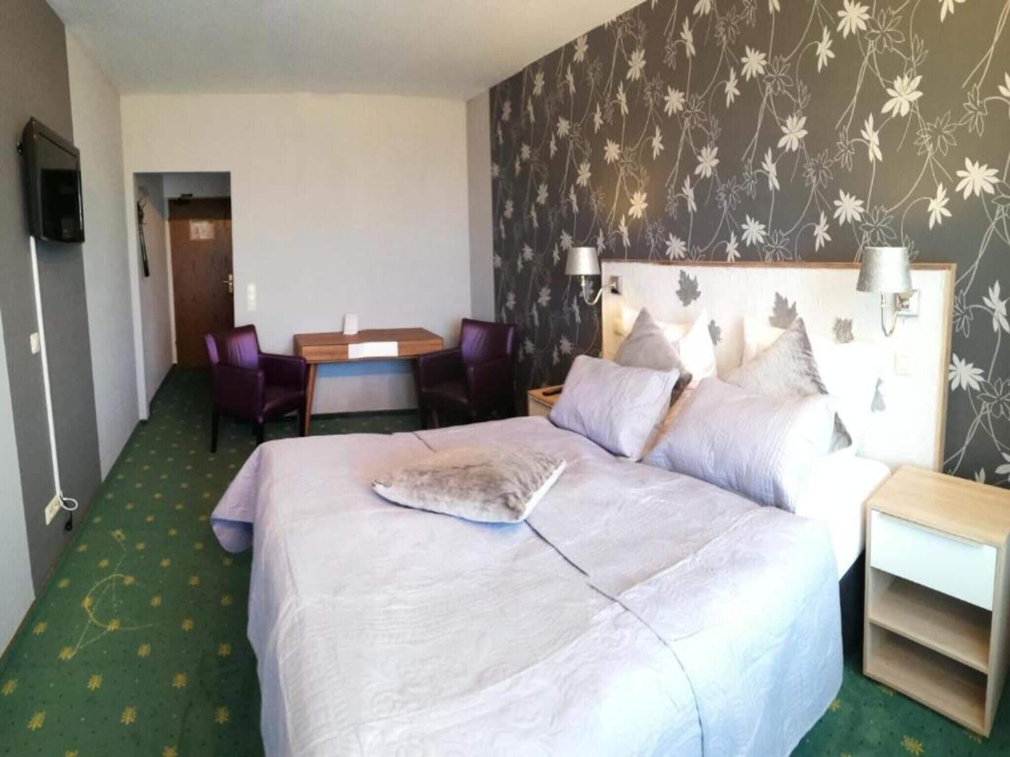 A hotel room with a bed, a desk, two chairs, a nightstand, and a wall-mounted TV.