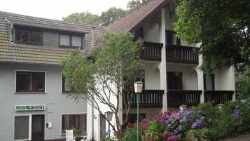 A two-story building with a balcony, a sign indicating a room number, and a small garden with flowers.