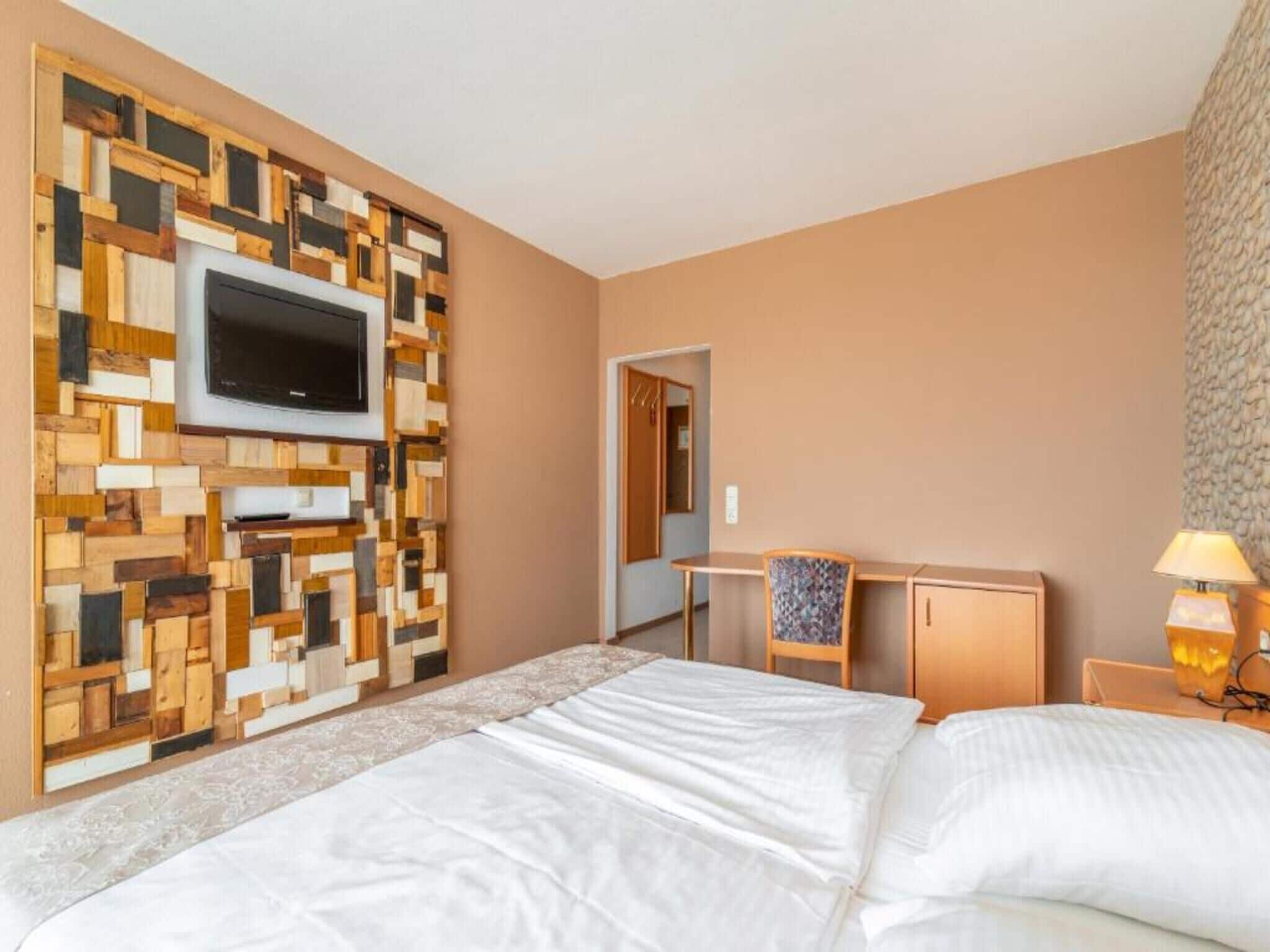 Habitación de hotel con cama, escritorio, silla, televisor montado en una pared de madera y lámpara.