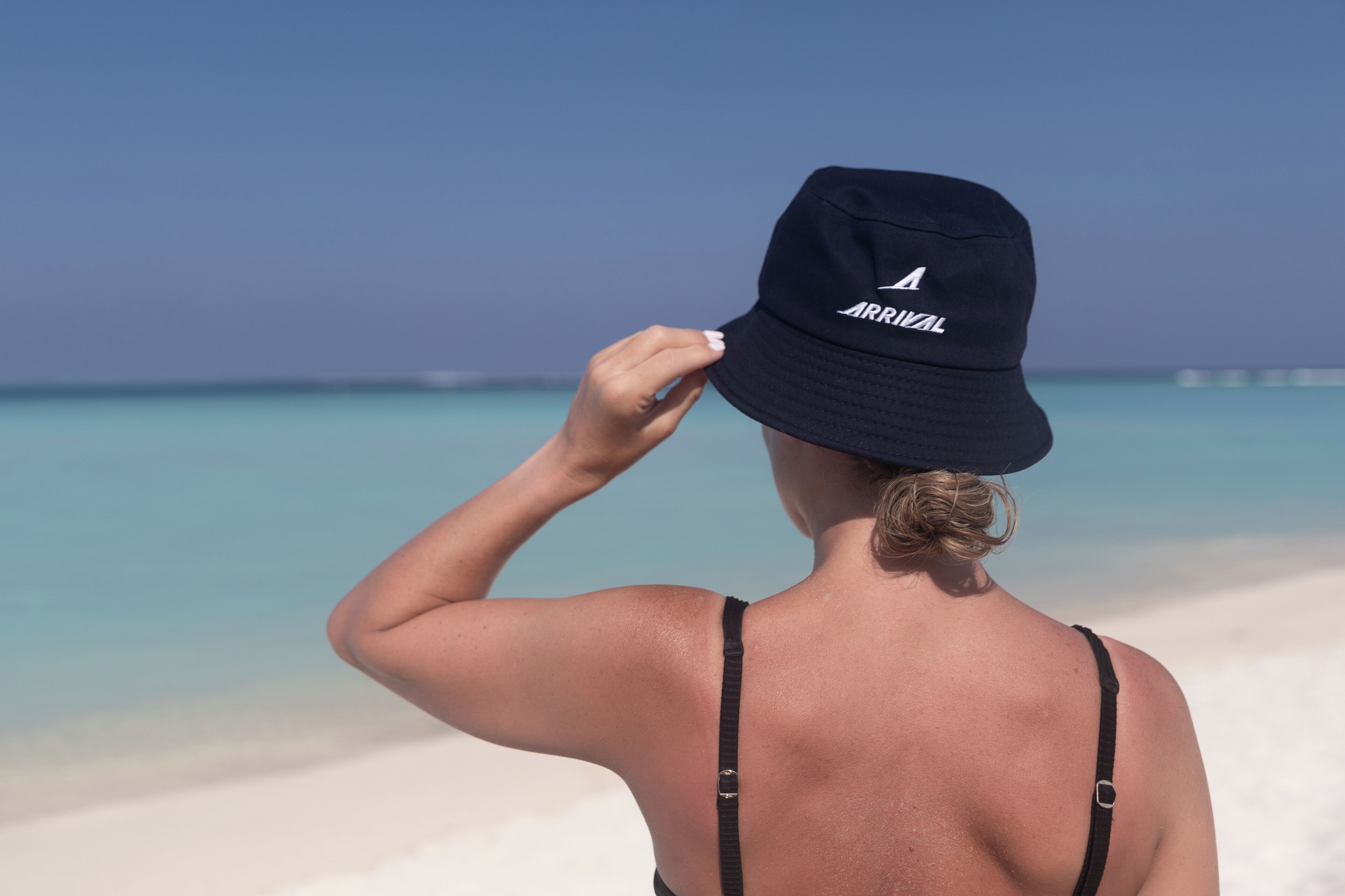A person wearing a navy blue bucket hat with "ARRIVAL" on it, standing on a beach with white sand and clear blue water.