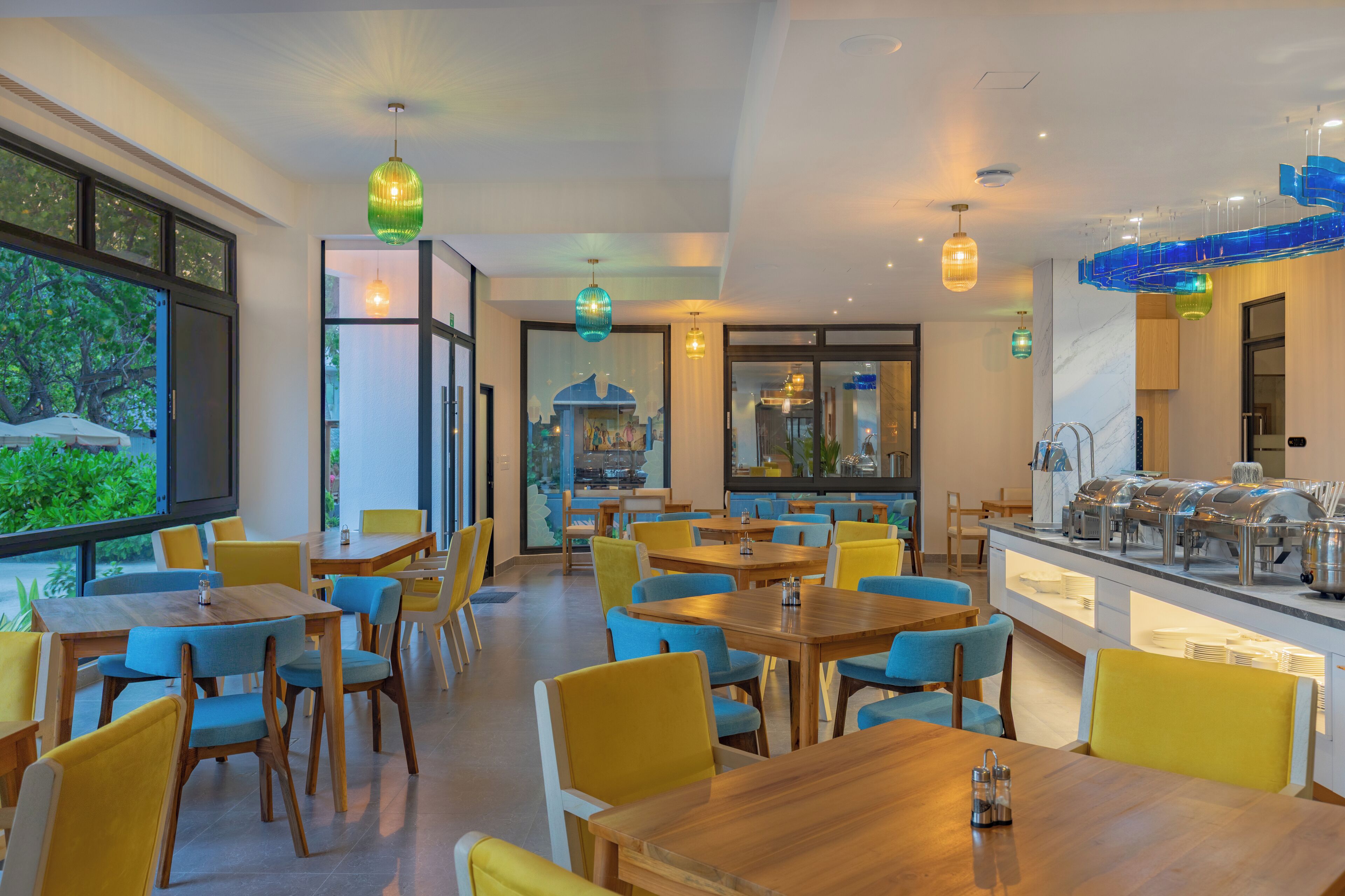 Un comedor moderno con mesas de madera y sillas amarillas, una barra de buffet y ventanas amplias con vistas a la vegetación.