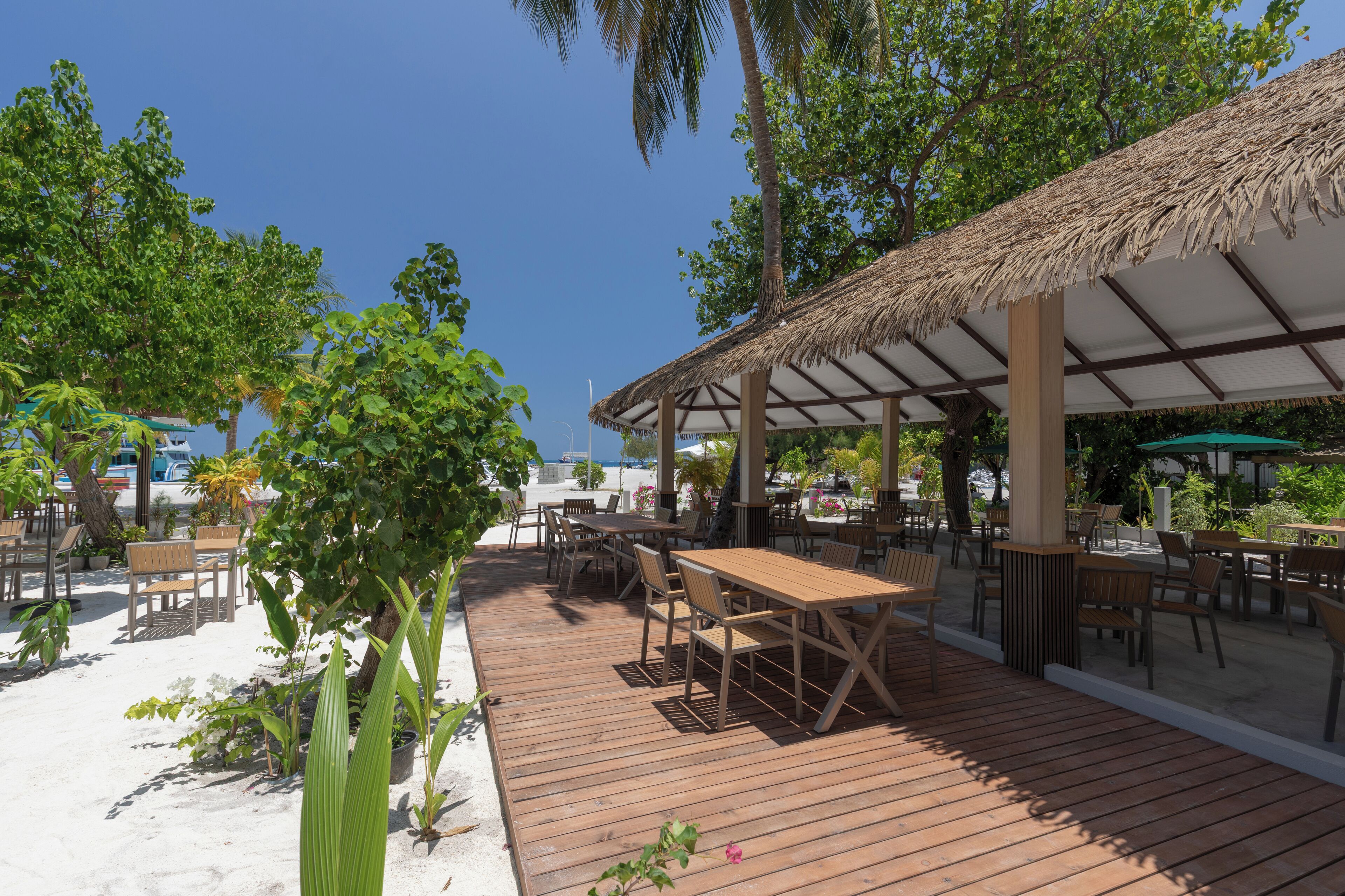 A wooden deck with tables and chairs, surrounded by palm trees and a thatched-roof structure.
