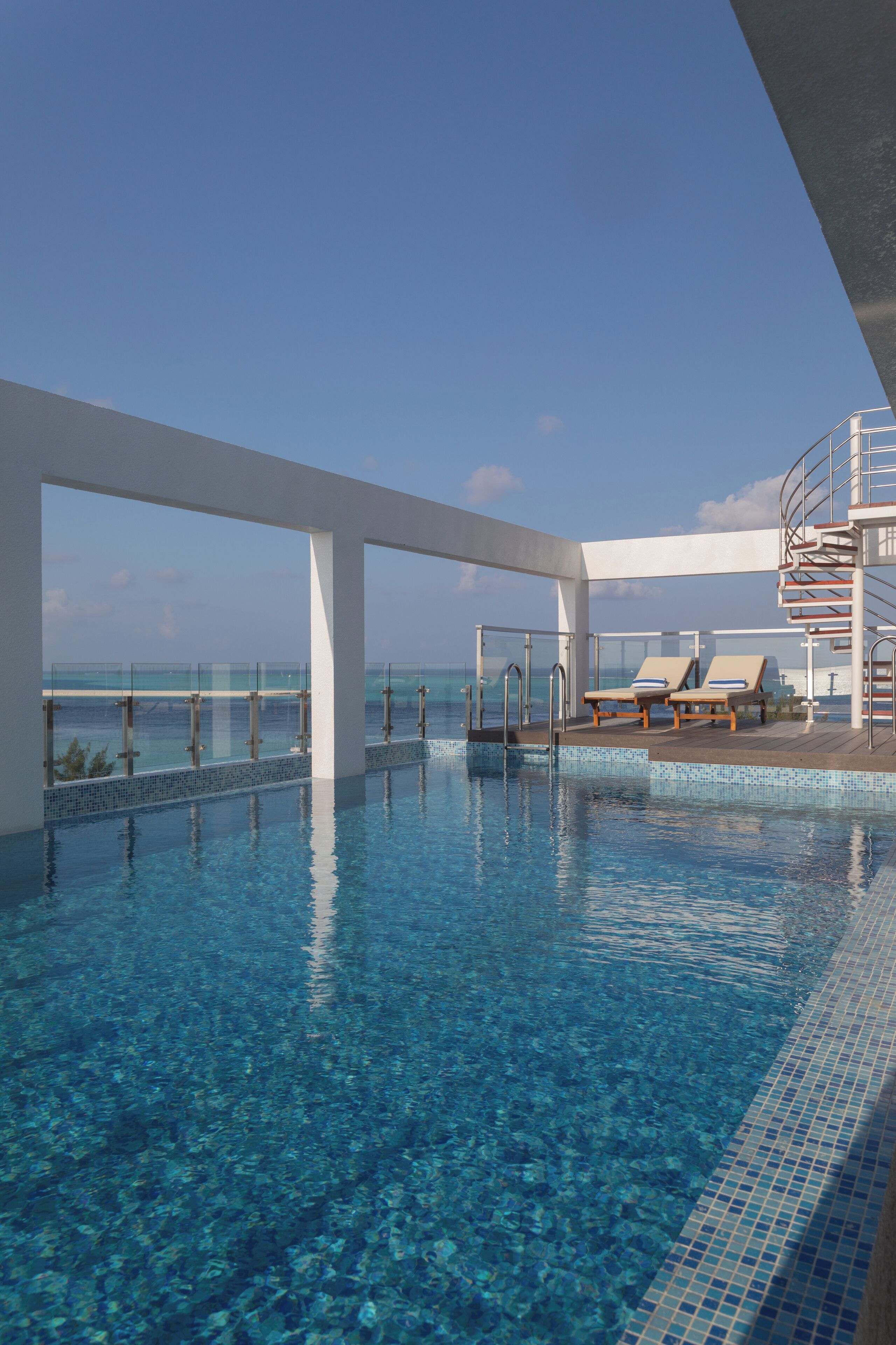 A clear blue pool with a glass balustrade, lounge chairs, and a staircase leading to an upper deck with a view of the ocean.