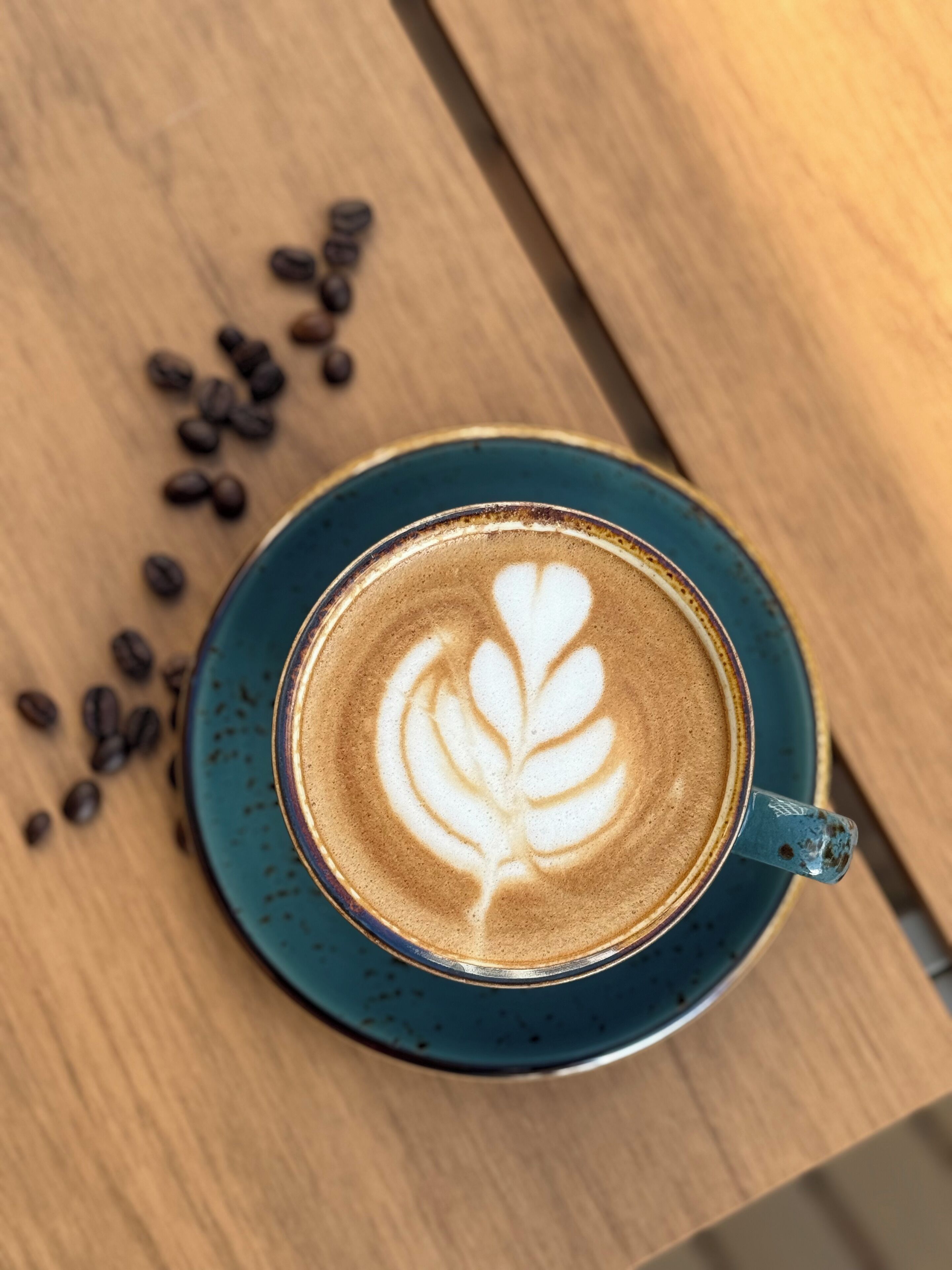 Una taza de café con decoración de latte sobre un platito azul, colocada sobre una mesa de madera con granos de café esparcidos alrededor.