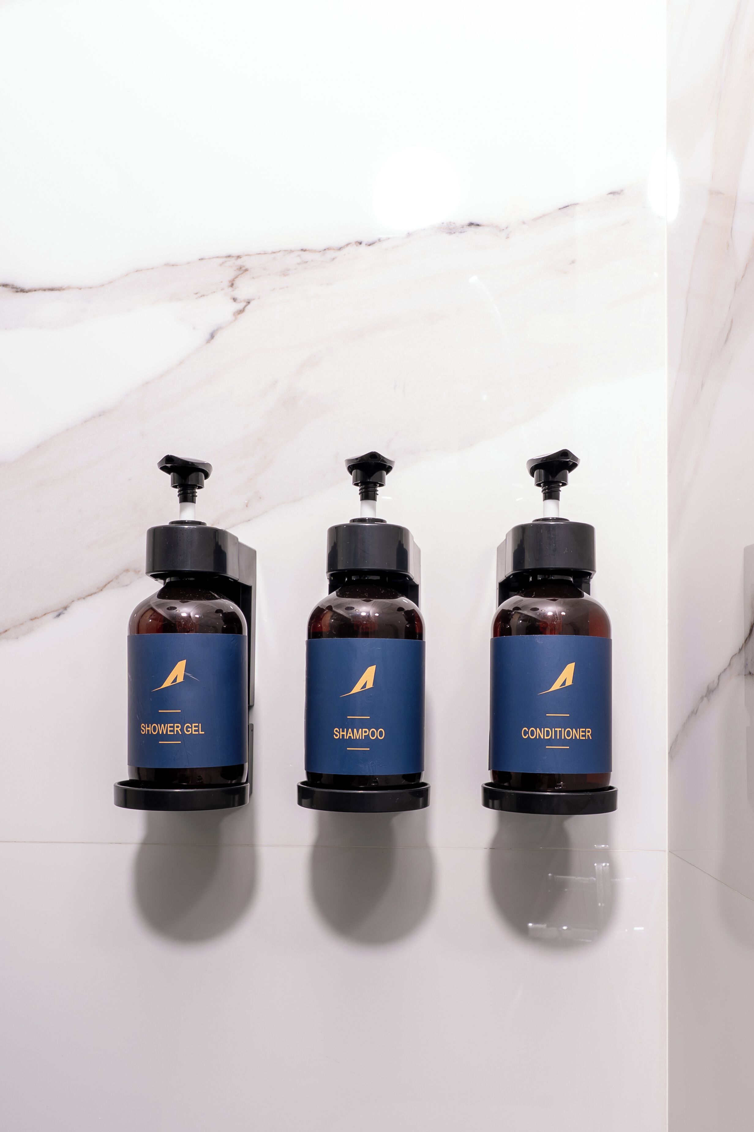 Trois bouteilles de gel douche, de shampoing et d’après-shampoing sont posées sur un mur de marbre blanc.