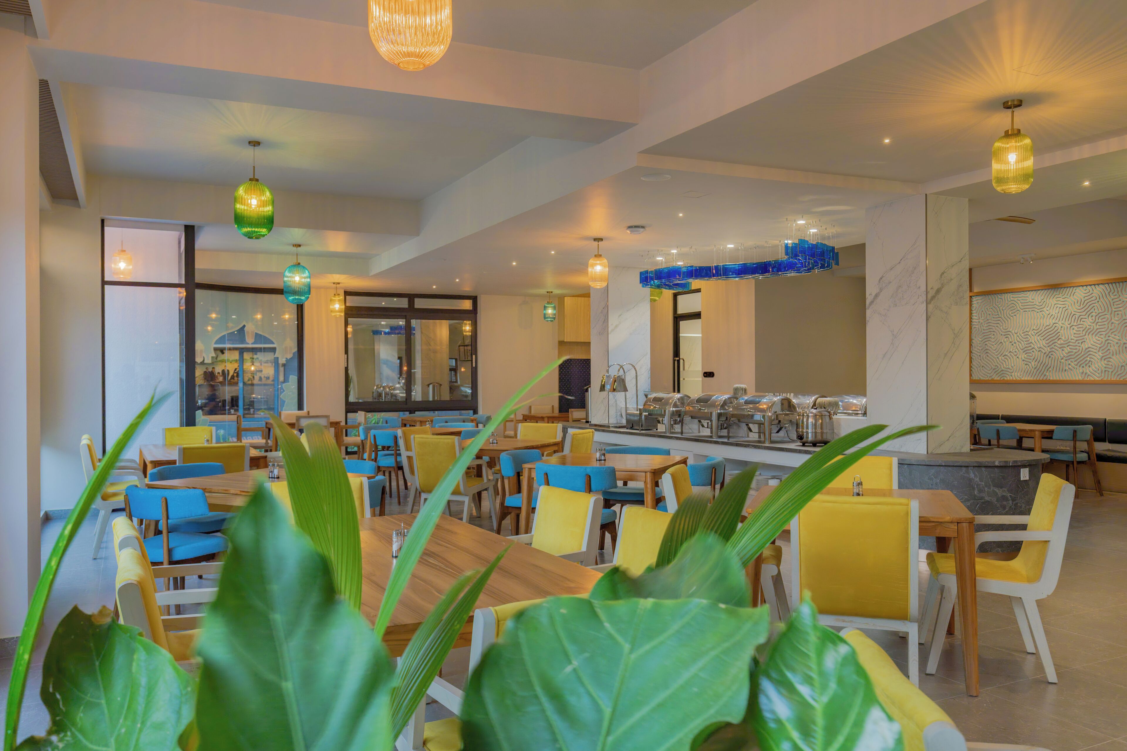 A modern restaurant with wooden tables and yellow chairs, large windows, and hanging green and yellow pendant lights.