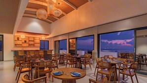 Un amplio comedor con varias mesas y sillas redondas, grandes ventanales con vistas al atardecer y un diseño de techo moderno.