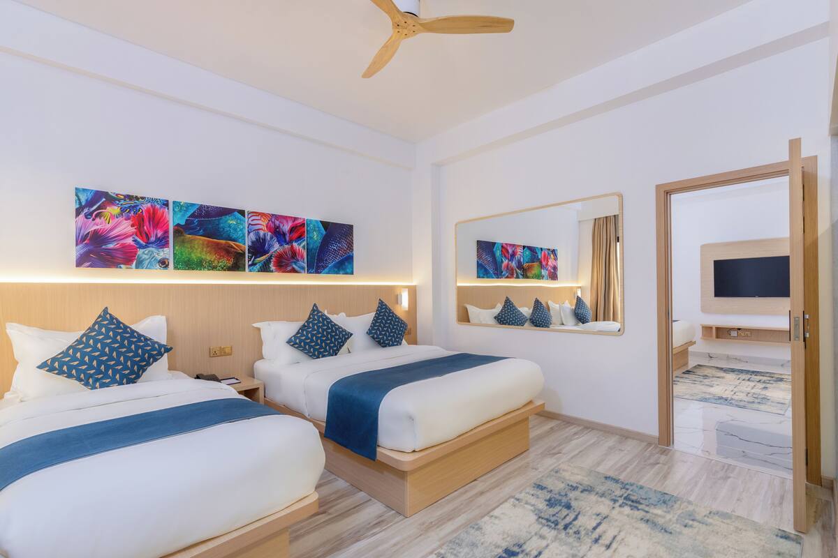 A hotel room with two beds, a ceiling fan, artwork on the walls, a TV, and a mirror.
