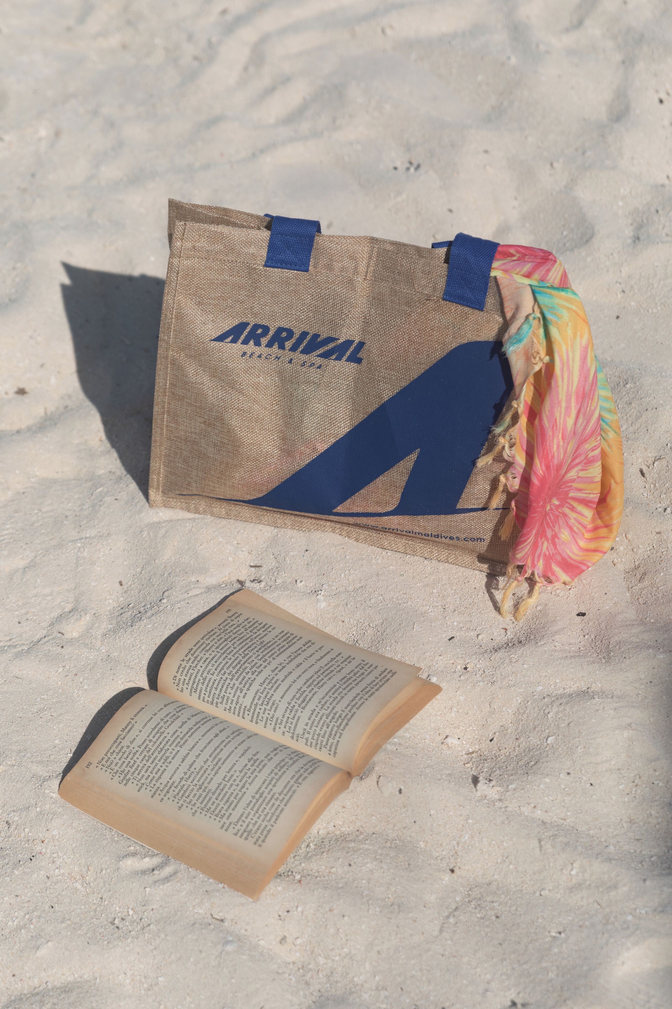 A burlap bag with a blue 'ARRIVAL' logo on a sandy surface next to an open book.