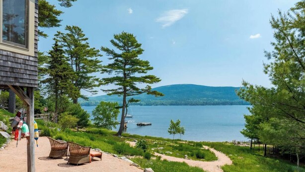 A lakeside cabin with a deck overlooking the water, surrounded by trees and a clear view of the lake and distant hills.