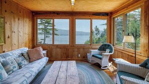 A wooden cabin with a large window overlooking a lake, furnished with a sofa, chairs, and a wooden table.