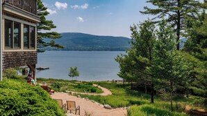A lakeside cabin with a deck overlooking the water, surrounded by greenery and a clear view of the lake and distant hills.