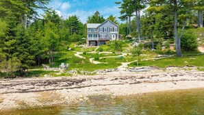 A two-story house with a balcony, surrounded by a forested area and a sandy beach.
