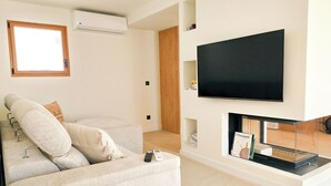 A modern living room with a sofa, a flat-screen TV mounted on the wall, and a fireplace with a magazine rack below. - Two Bedroom Villa (Porches)